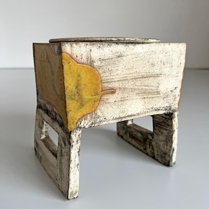 May include: A handcrafted ceramic box with a lid, featuring a rectangular shape and a leaf design. The box is supported by a base with cut-out details. The color palette includes cream, yellow, and brown tones, creating a rustic aesthetic.