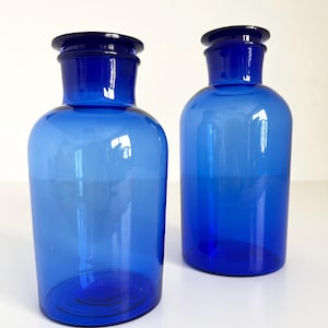 May include: Two cobalt blue glass bottles with a rounded body and a wide mouth. The bottles have a dark blue glass stopper.