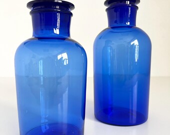 May include: Two cobalt blue glass bottles with a rounded body and a wide mouth. The bottles have a dark blue glass stopper.
