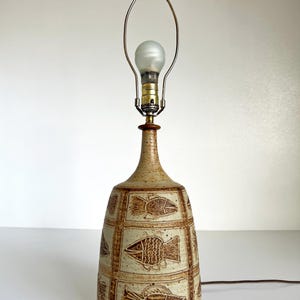 May include: A ceramic table lamp with a beige and brown fish design. The lamp has a bulb and a metal shade holder. The lamp is approximately 15 inches tall.