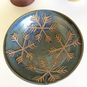 May include: A blue ceramic bowl with a snowflake design. The bowl is glazed in a dark blue color with a light blue glaze on the rim. The snowflakes are a light brown color and are raised from the surface of the bowl.