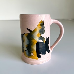 May include: A light pink ceramic mug featuring a hand-painted calico cat design in shades of orange, black, and white, alongside a solid black cat silhouette. The mug has a smooth, glossy finish and a curved handle on the right side.