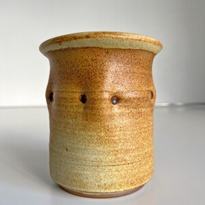 May include: A handmade ceramic jar with a cylindrical shape and a slightly flared rim. The jar features a speckled, earthy color palette of tan and brown, with a series of small, evenly spaced holes around the middle. The jar is a decorative item.