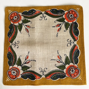 May include: A square linen napkin with a mustard yellow border and a white center. The border features a repeating floral pattern in shades of red, orange, green, and blue.