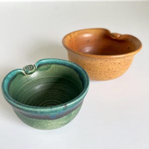 May include: Two handmade ceramic bowls, one green with a blue rim and one brown with a speckled glaze. Both bowls have a unique, asymmetrical shape with a small handle on the side.
