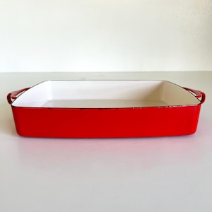 May include: A rectangular red baking dish with a white interior and black trim. The dish has two red handles on either side. This enamelware dish is perfect for baking casseroles, roasting vegetables, or making desserts.