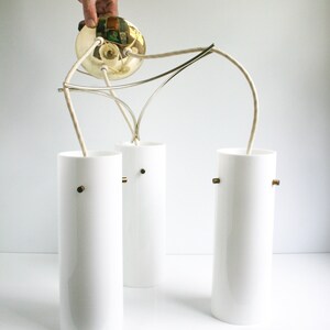 May include: A three-light pendant light fixture with a brass canopy and white cylindrical glass shades. The fixture has a modern design with a simple, elegant look.