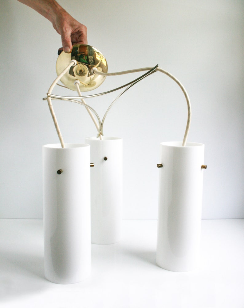 May include: A three-light pendant light fixture with a brass canopy and white cylindrical glass shades. The fixture has a modern design with a simple, elegant look.
