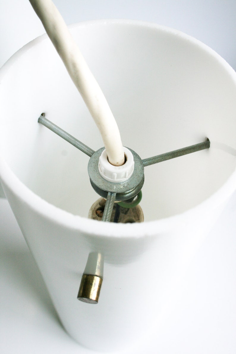May include: Close-up of a white ceramic lampshade with a metal socket and wiring attached. The socket has three screws and a white plastic cover.