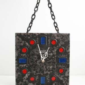 May include: A black ceramic clock with red and blue geometric shapes and a white chain hanging from the top. The clock face has a white background with black hands and the text "LENZLE ANTIQUE" in white.