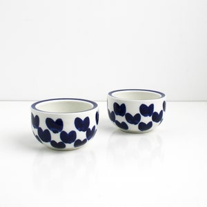 May include: Two white ceramic bowls with a blue and white heart pattern. The bowls are small and round.