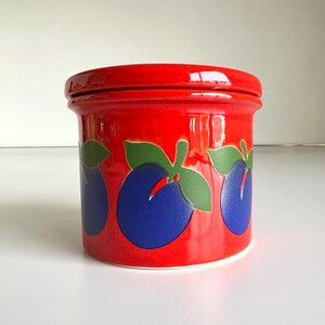 May include: A cylindrical ceramic canister with a glossy red glaze, featuring a repeating pattern of dark blue plums with green leaves around the center. The canister includes a matching red lid that sits flush with the top rim of the container.