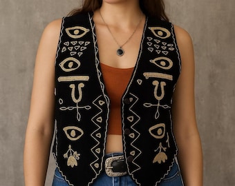 May include: Black vest with gold embroidered geometric and eye designs. The vest has a button-up front and a decorative silver buckle on the belt. The vest is worn over a brown top and blue jeans.