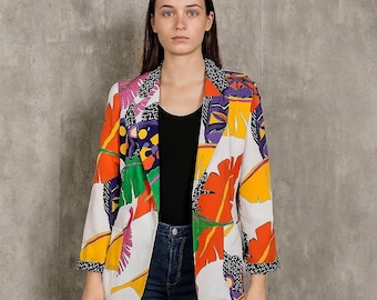 May include: A white blazer with a vibrant, colorful pattern featuring orange, yellow, purple, and green leaf-like shapes. The blazer has a black and white patterned trim on the collar and cuffs. The model is wearing a black top and blue jeans.