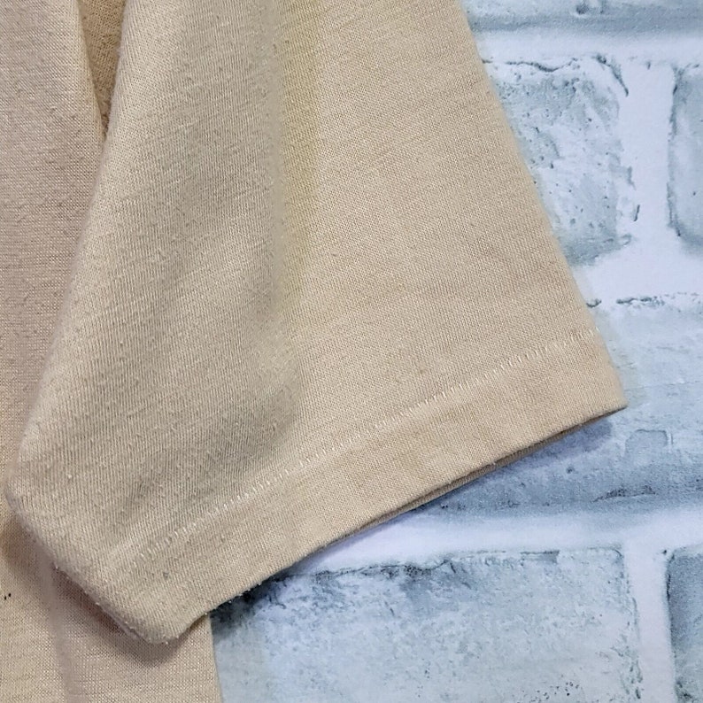 May include: Close-up of the beige fabric sleeve of a shirt. The fabric has a textured, slightly rough surface. The sleeve is folded over and the seam is visible.