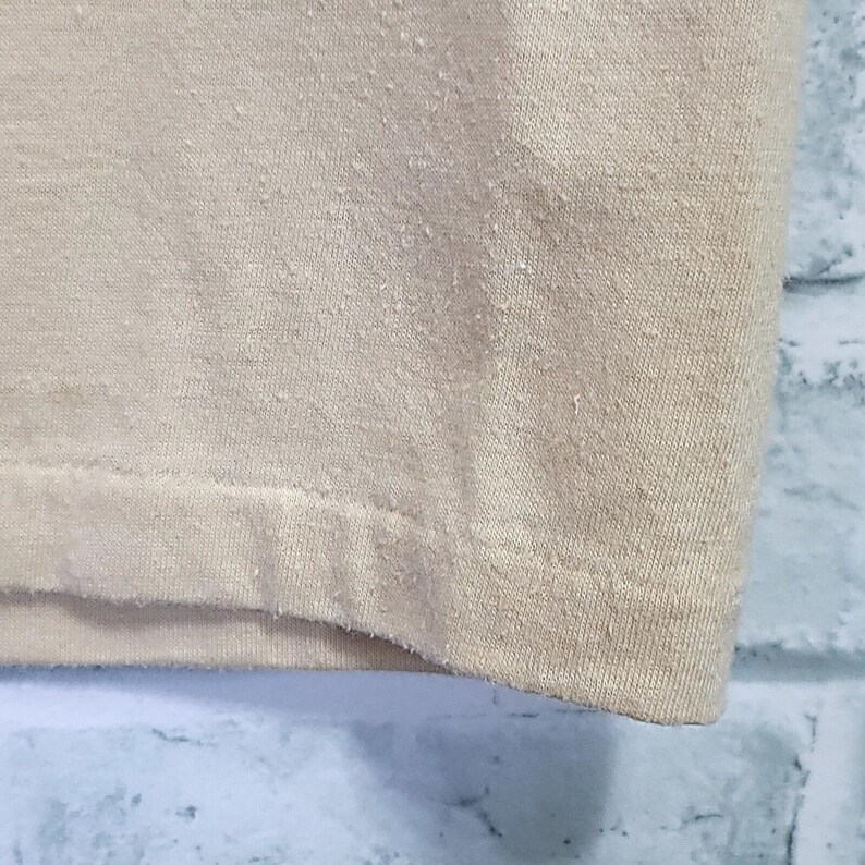 May include: Close-up of the bottom hem of a beige t-shirt. The fabric is a soft, knit material.