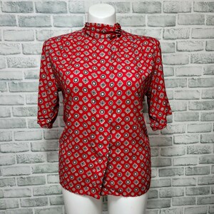 May include: A red short-sleeved button-up shirt with a white and black geometric pattern. The shirt has a high collar and a fitted silhouette.