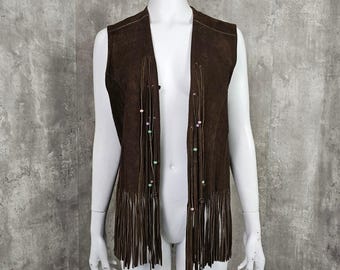 May include: A brown suede vest with a fringed hem and beaded accents. The open-front vest features long fringe along the bottom and decorative beaded strands. The vest is displayed on a mannequin against a neutral background.
