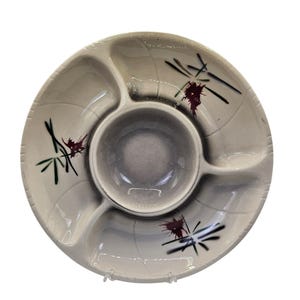 Puede incluir: Una fuente redonda y dividida con un cuenco central para salsas. La fuente es de color blanquecino con un centro y cuenco para salsas grises. Presenta un diseño floral estilizado en negro, verde y granate. La fuente es de cerámica.