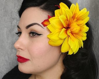 May include: A woman with dark hair and red lipstick, wearing a large yellow flower hair accessory. The flower has a vibrant yellow color with orange and red accents. The woman is wearing black clothing.