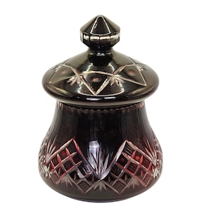 Op de afbeelding: Een donkerrode glazen pot met deksel, voorzien van ingewikkelde geslepen glasontwerpen. De pot heeft een afgeronde basis en een conisch deksel. De geslepen glaspatronen omvatten ruit- en sterrenmotieven, wat een decoratieve uitstraling geeft.