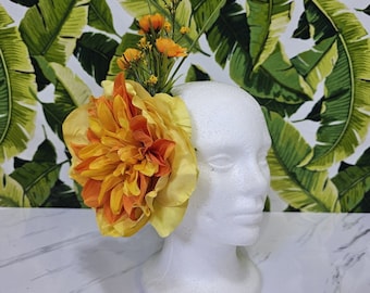 May include: A large, bright orange and yellow flower with a cluster of smaller yellow flowers on a white mannequin head. The background is a green and yellow tropical leaf pattern.