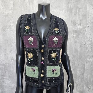 May include: Black velvet vest with floral embroidery. The vest features a V-neck, button closure, and a patchwork design with embroidered flowers in gold, white, and purple. The vest is displayed on a black mannequin.