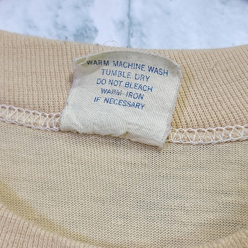 May include: A white care tag with blue text that reads "Warm Machine Wash, Tumble Dry, Do Not Bleach, Warm Iron If Necessary."