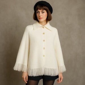 Może przedstawiać: Kremowy płaszcz typu peleryna z kołnierzem i frędzlami. Płaszcz ma złote guziki z przodu. Modelka ma na sobie czarny beret i czarne szorty. Płaszcz jest wykonany z teksturowanej tkaniny.