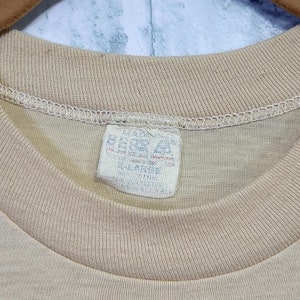 May include: A beige t-shirt with a white tag that reads "Made in USA, 100% Cotton, Machine Wash, Tumble Dry Low, Do Not Bleach".