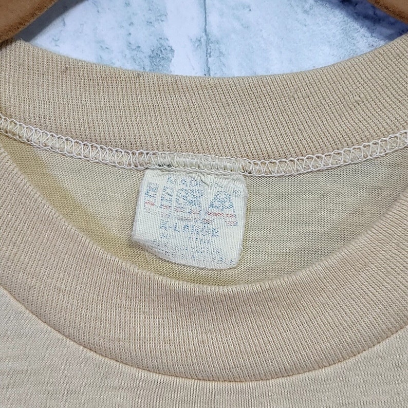 May include: A beige t-shirt with a white tag that reads "Made in USA, 100% Cotton, Machine Wash, Tumble Dry Low, Do Not Bleach".