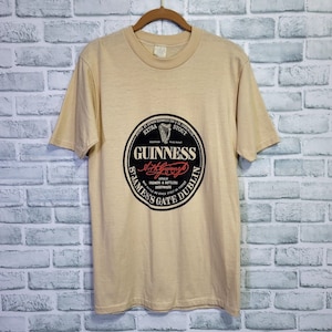 May include: A tan t-shirt with a black and white Guinness logo. The logo features a harp, the words "Extra Stout", and the text "Brewers & Bottlers Everywhere".
