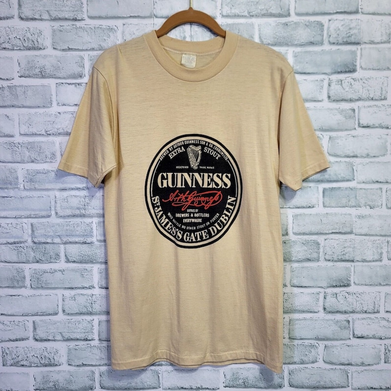 May include: A tan t-shirt with a black and white Guinness logo. The logo features a harp, the words "Extra Stout", and the text "Brewers & Bottlers Everywhere".