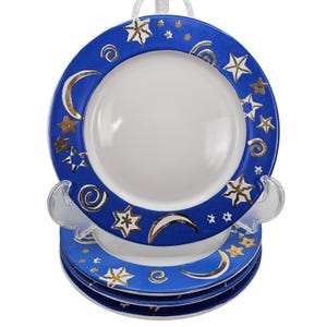 May include: A set of four ceramic plates with a celestial design. Each plate has a white centre and a blue border decorated with gold stars, crescent moons, and spiral accents. The plates are stacked on a clear plastic stand.
