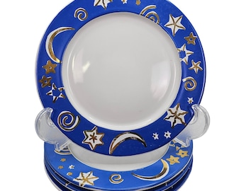 May include: Set of four ceramic plates with a celestial design. Each plate features a white center and a blue border adorned with gold stars, crescent moons, and spiral accents. The plates are stacked on a clear plastic stand.