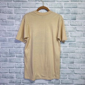 May include: A light beige short-sleeved t-shirt hanging on a wooden hanger against a white brick wall background.