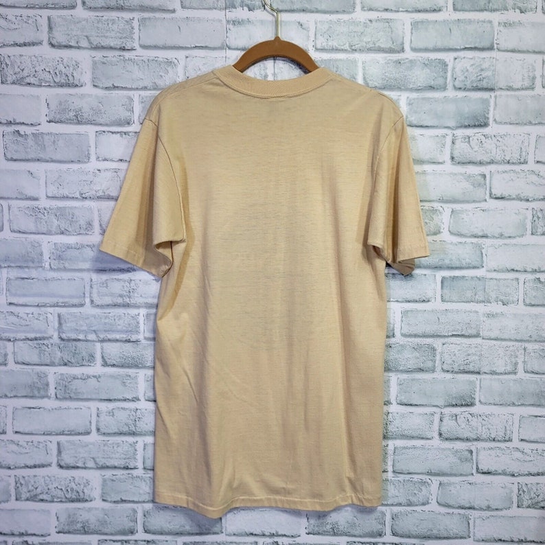 May include: A light beige short-sleeved t-shirt hanging on a wooden hanger against a white brick wall background.
