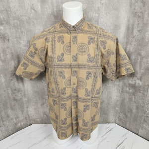 May include: Tan short-sleeve button-down shirt with a repeating navy blue paisley pattern. The shirt is displayed on a white mannequin. The pattern is arranged in square blocks across the shirt.