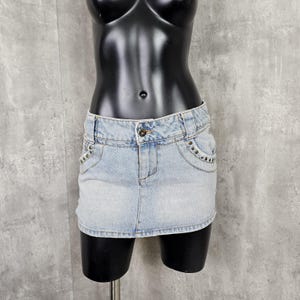 May include: Light blue denim mini skirt with a button closure and pockets. The skirt has decorative rivets along the pocket edges. The garment is displayed on a black mannequin.