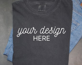 May include: A dark gray t-shirt with the text "your design HERE" printed in white on the front. The t-shirt is folded and laid on top of a pair of blue jeans.
