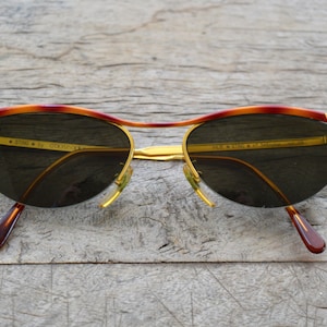 May include: Gold-tone sunglasses with a tortoiseshell frame and dark green lenses. The sunglasses have the text "STAR BY COCCINELLI" on the left lens and "MADE IN ITALY" on the right lens.