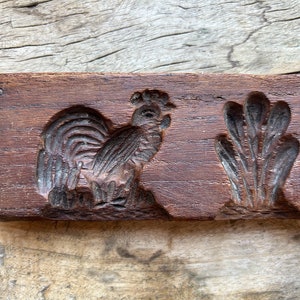 May include: A vintage wooden cookie mold with two designs: a rooster and a cluster of leaves. The mold is dark brown and has a worn, rustic appearance.