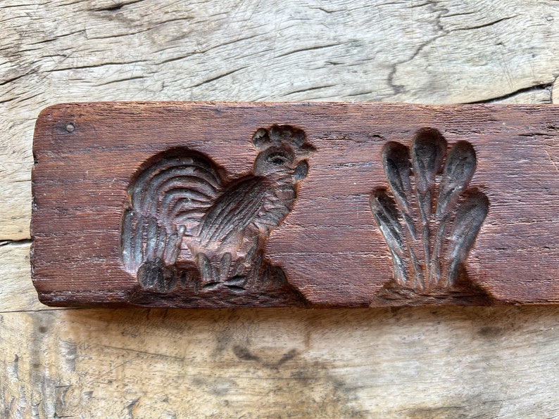 May include: A vintage wooden cookie mold with two designs: a rooster and a cluster of leaves. The mold is dark brown and has a worn, rustic appearance.