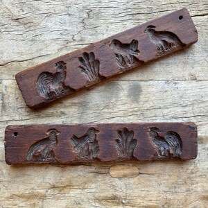May include: Two wooden cookie cutters with carved designs of roosters, chickens, and wheat stalks. The cutters are made of dark brown wood and have a rustic appearance.