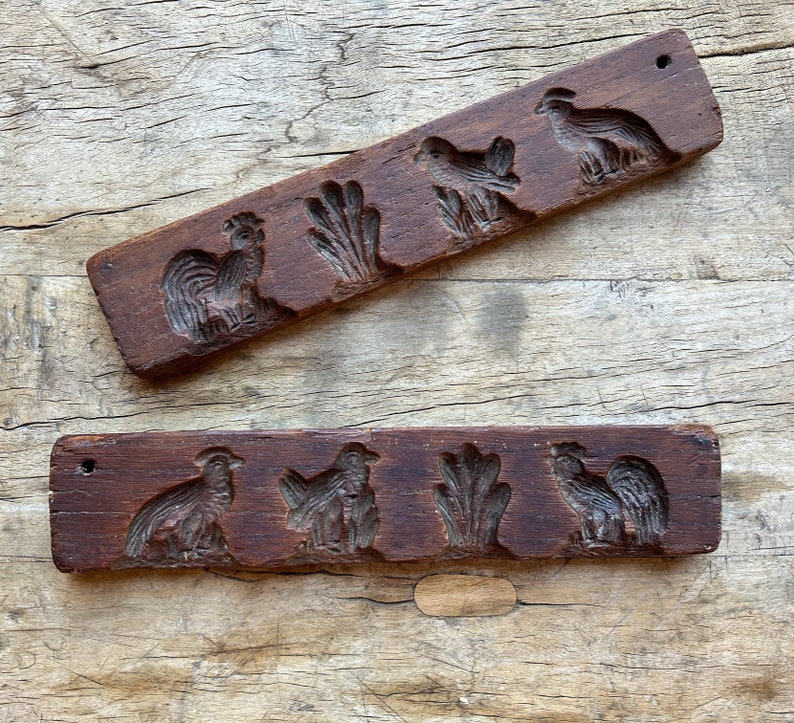 May include: Two wooden cookie cutters with carved designs of roosters, chickens, and wheat stalks. The cutters are made of dark brown wood and have a rustic appearance.