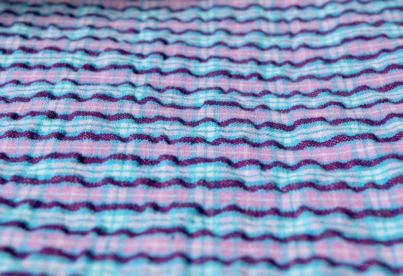 K&ouml;nnte beinhalten: Nahaufnahme eines strukturierten Stoffes mit einem wellenf&ouml;rmigen Muster. Der Stoff ist blau, lila und wei&szlig;.