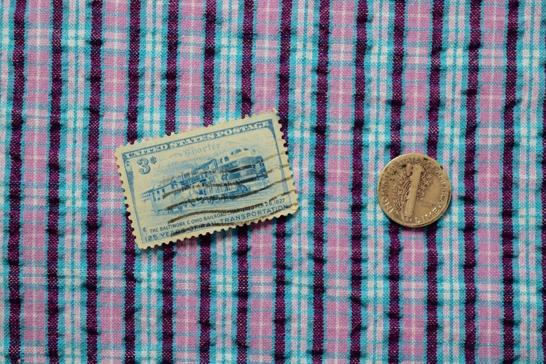 K&ouml;nnte beinhalten: Eine blaue und wei&szlig;e Briefmarke der Vereinigten Staaten mit einem Zug darauf und dem Text "3&cent;" und "The Baltimore & Ohio Railroad 125 Years of Transportation 1827-1952". Eine Silberm&uuml;nze mit dem Text "Liberty" und "In God We Trust" liegt auf einem rosa-blauen karierten Stoff.