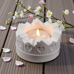 Könnte beinhalten: Ein weißer Kerzenhalter mit herzförmigem Design. Der Kerzenhalter hat eine kronenartige Oberseite und eine brennende Kerze im Inneren. Rosa und weiße Blüten und Blütenblätter sind um den Kerzenhalter auf einer Holzoberfläche verstreut.