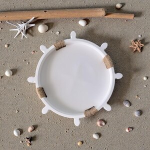 Könnte beinhalten: Ein weißes Serviertablett in Form eines Schiffsrads mit Seilakzenten. Das Tablett ist von Muscheln, Seesternen und Treibholz auf sandigem Untergrund umgeben. Ein dekorativer Artikel für die Küstendekoration.