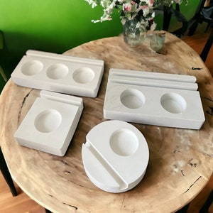 May include: Four white ceramic coasters with different shapes and sizes. The coasters are on a wooden table with a vase of white flowers in the background.